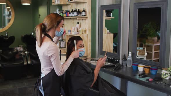 Female hairdresser and female customer wearing face masks looking at smartphone at hair salon alt
