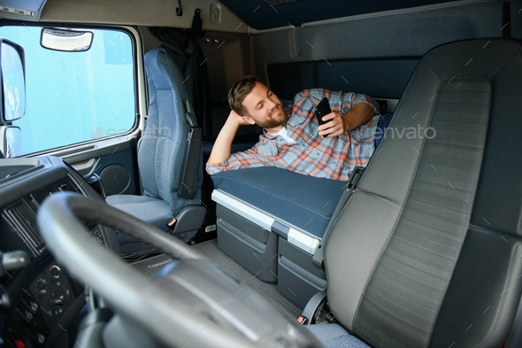 Truck driver lying on bed in his truck cabin behind driver seat and holding phone chatting