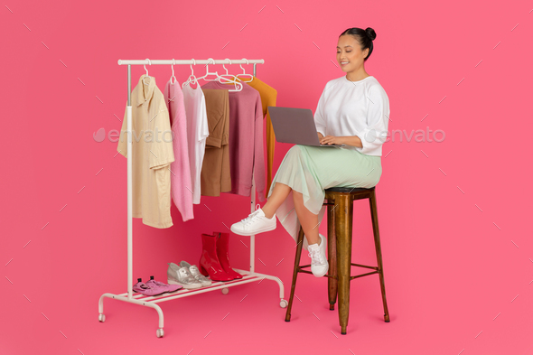 Beautiful asian woman sitting with laptop near clothing rack, managing online boutique