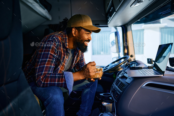Happy black driver using laptop while eating lunch in his truck cabin.
