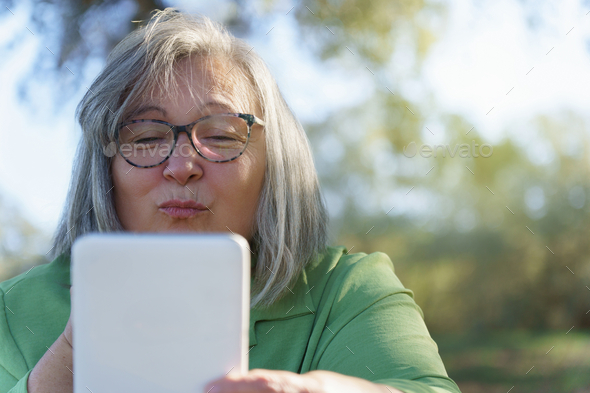 mature white-haired woman with glasses blowing a kiss to her digital tablet