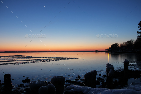 Winter sunset landscape with frozen body of water and seashore silhouettes