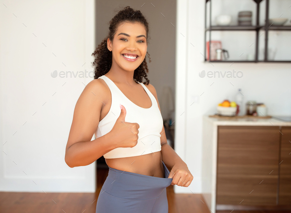 woman gesturing thumbs up pulling waist of fitness pants indoors