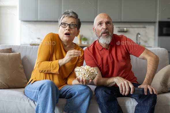 Shocked elderly couple sitting on couch with bowl of popcorn, watching tv