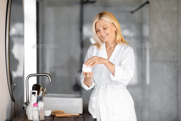 Smiling senior woman in white bathrobe opening jar of face cream