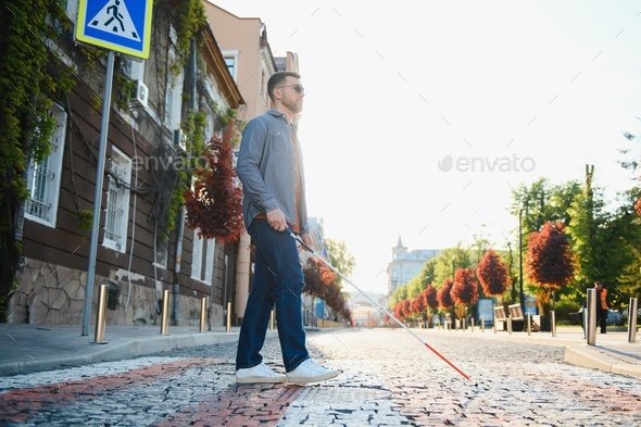 Young blind man with white cane walking across the street in city