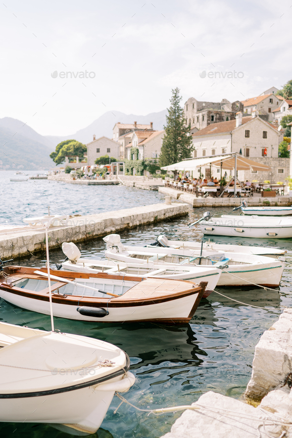 Row of fishing boats moored at the Perast pier against the backdrop of  ancient stone houses at the Stock Photo by Nadtochii