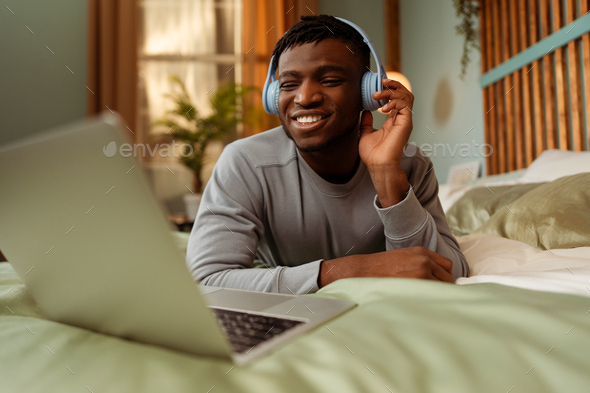 Happy smiling African American man wearing headphones lying on bed using laptop having video call