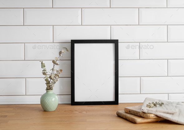 A black frame with a blank canvas against a white tile wall and on a wooden tabletop