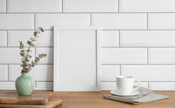 A frame with a blank canvas against a white tile wall and on a wooden tabletop with vase and cup