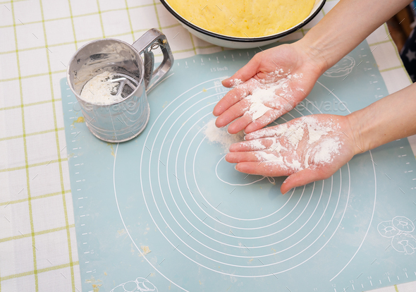 The image shows hands covered in flour over a silicone baking mat. A sifter and mixing bowl are