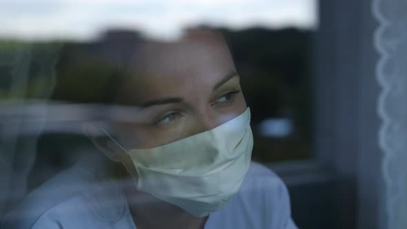 Young Woman Wears a Mask To Protect Herself From Covid19 While Looking Out the Window