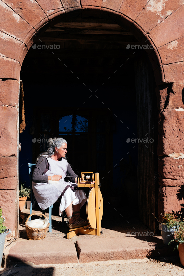 Elderly woman working on wooden spinning wheel in old building