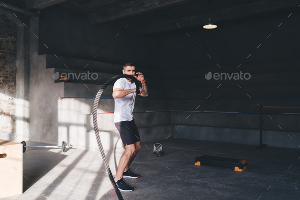 Athletic man pulling battle rope in workout in gym Stock Photo by