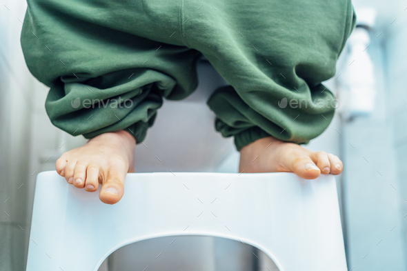adult male sitting on white toilet with his pants down and feet are standing on toilet foot Stool