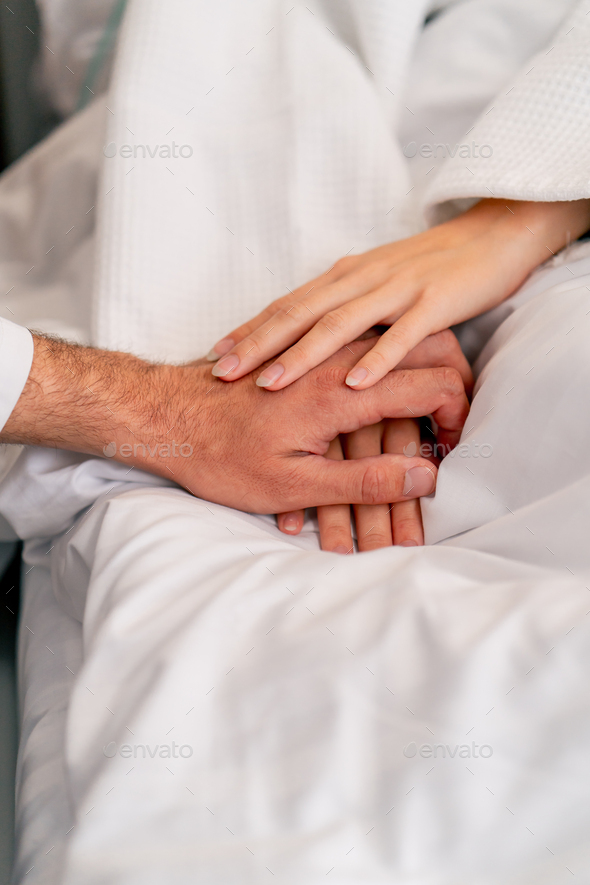 a male doctor\'s hand holding a female patient\'s hand on a bed in a hospital ward for moral support