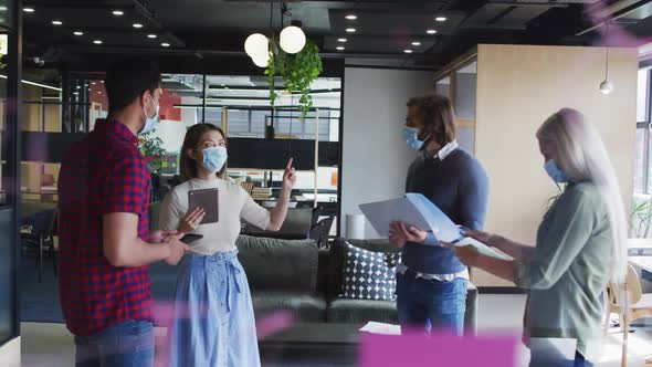 Diverse business people wearing masks standing using laptop and goign through paperwork in office alt