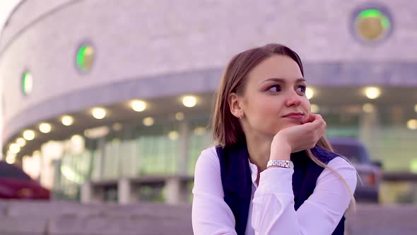 A Young Girl in a White Shirt and Vest, Sitting Outside in the Evening, Waiting for a Meeting.