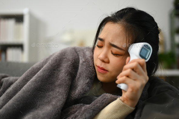 Young woman measuring fever with infrared thermometer and touching forehead.