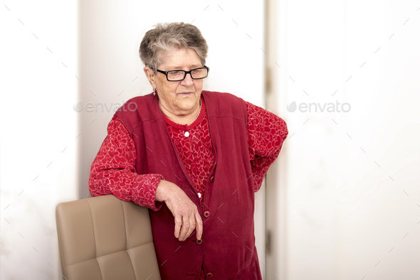 An old woman 70+ years old holds her hand behind her back, leans her elbows on a chair, looking tire
