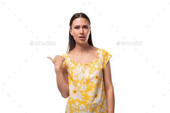 Caucasian young woman with straight black hair dressed in a yellow sundress points with her hand to