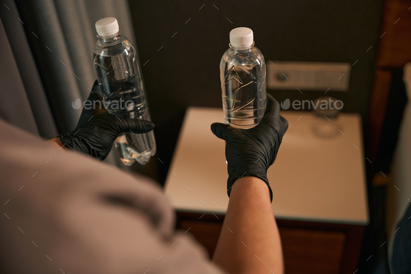 Maid in disposable gloves putting beverages on nightstand in hotel room