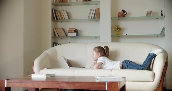 A Little Girl Lies at Home on the Couch and Looks Into a Laptop alt