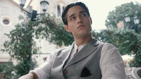 Attractive young man in a suit on a summer terrace drinking soda and waiting for someone alt