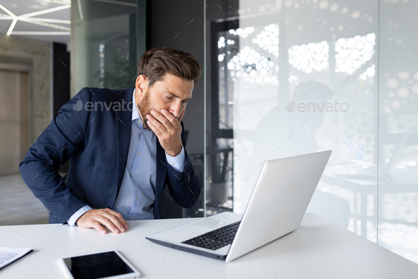 A young man businessman sits in the office at the work table and covers his mouth from nausea, he