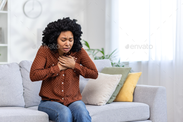 Worried young woman sitting on couch at home feeling chest pain, possible heart attack, stress or