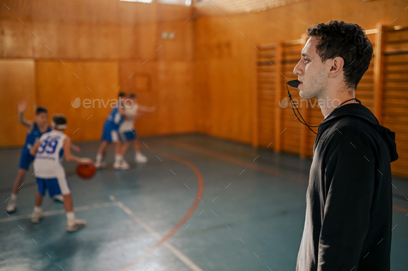 Side view of a trainer on court using whistle while boys practicing basketball.
