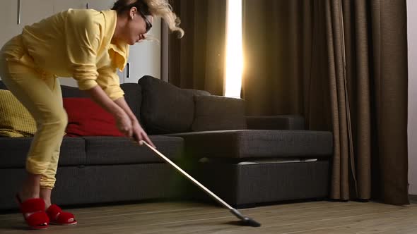Happy Blonde Woman in a Yellow Hoodie with a Mop Cleaning the House and Dancing alt