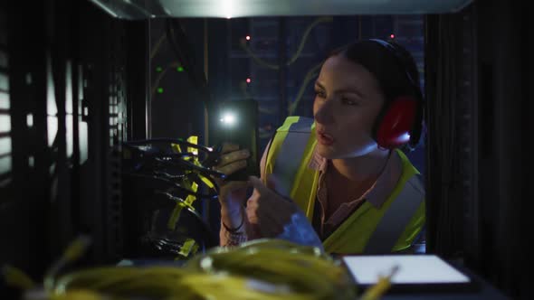 Caucasian female it technician wearing headphones and using torch checking computer server alt