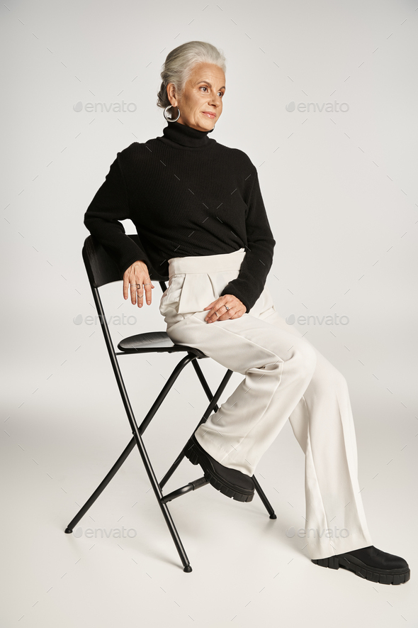 elegant middle aged business woman in smart casual attire and hoop earrings sitting on chair on grey