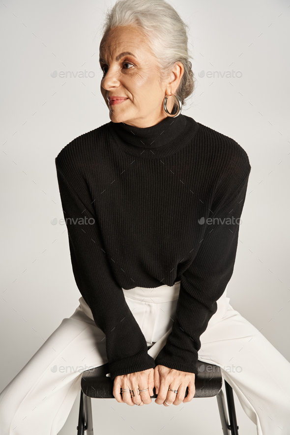 pleased middle aged business woman in smart casual attire and hoop earrings sitting on chair on grey