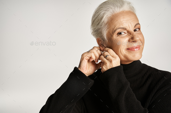happy middle aged business woman in elegant attire wearing hoop earrings on grey background