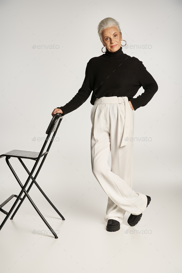 middle aged business woman in smart casual attire and hoop earrings standing near chair on grey