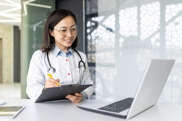 Asian doctor consulting patients online at clinic office with laptop and clipboard