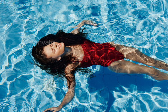 Happy woman swimming in pool in red swimsuit with loose long hair relaxed  with hands up in sunlight Stock Photo by shotprime