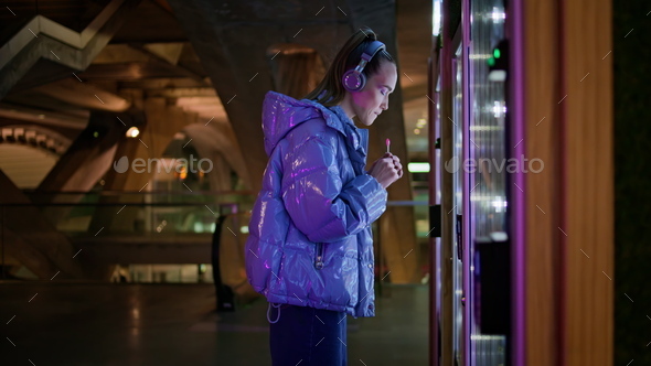Girl choosing snacks vending machine standing in subway. Woman in headphones.