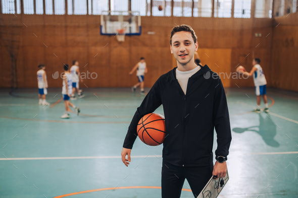 Portrait of a basketball coach standing with ball and clipboard on court.