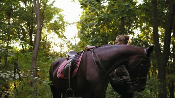 Female Equestrian Climbs on Horseback with Help of Stirrup During Sunny Day in the Forest alt