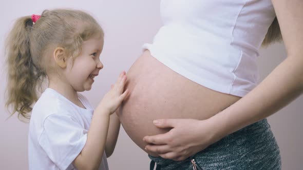 Happy Girl Plays with Pregnant Mother Belly Standing at Wall alt