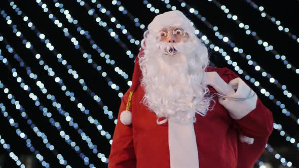 Santa Claus Showing Thumbs Up on the Background of Flashing Garlands Outdoors alt