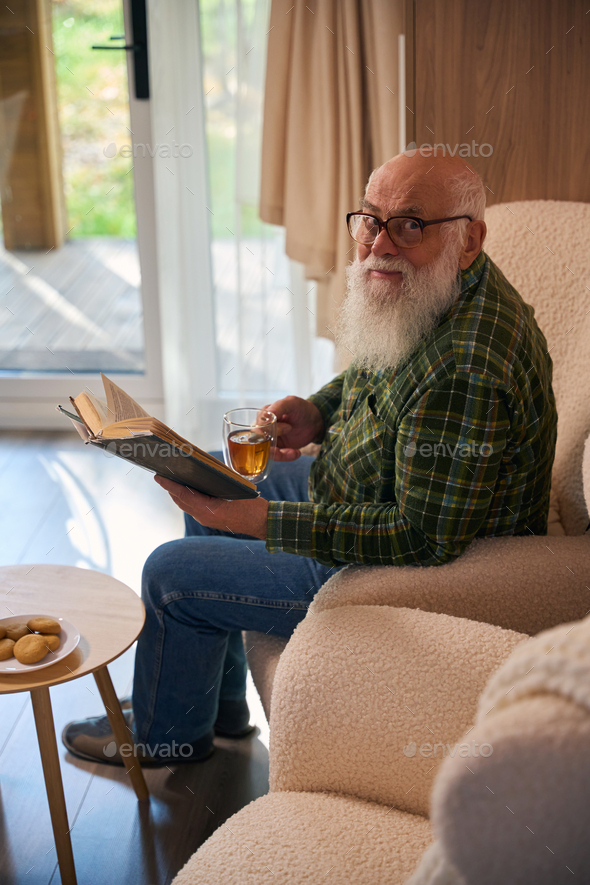 Old man with glasses settled down with book by French window
