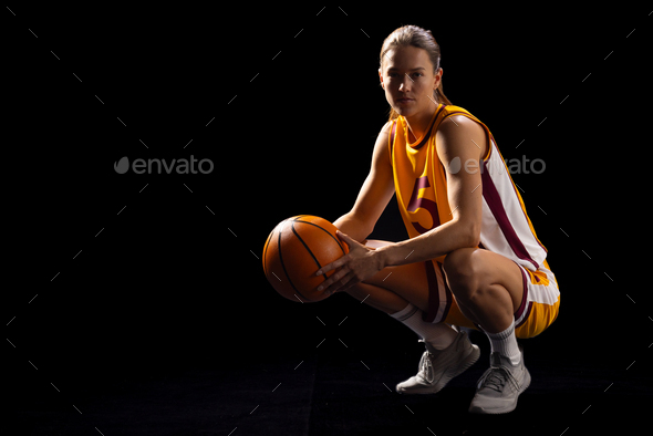 Young Caucasian female basketball player crouches in a basketball pose on a black background, with c