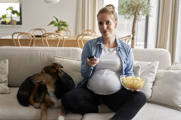 Pregnant woman watching TV and eating crisps with a dog
