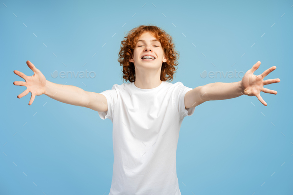 Red haired teenager male with braces, extending arms in welcoming hug pose on blue background