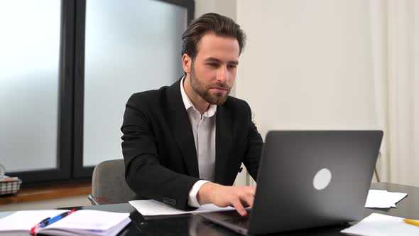 Crazy Excited Lucky Young Bearded Businessman Sitting at the Desk with Laptop Raised Fists Up alt