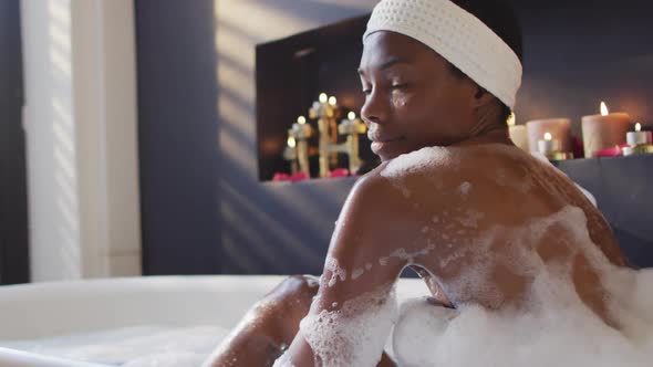 Smiling african american woman with towel taking bath and touching her skin in bathroom alt